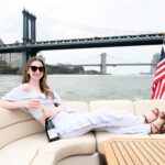 a woman with a bottle of champagne with the American flag and Brooklyn Bridge behind during boat rentals in NYC