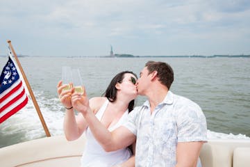 couple kissing on a yacht with an American flag and the Statue of Liberty in the background during a romantic cruise NYC