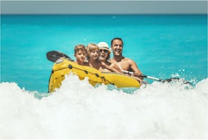 A happy family is enjoying a ride in a rubber kayak, and waves are splashing them in tropical ocean water during summer vacation.