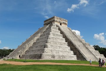 a large stone building with a grassy field with Chichen Itza in the background