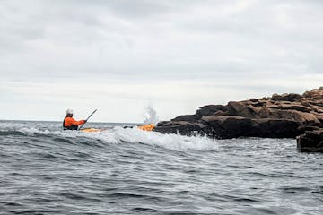 A sea kayaker paddling in roughwater near rocks and waves