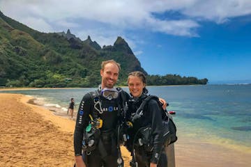 A couple scuba diving at Tunnels Beach, posing and smiling for a photo. The sky above is partly cloudy.