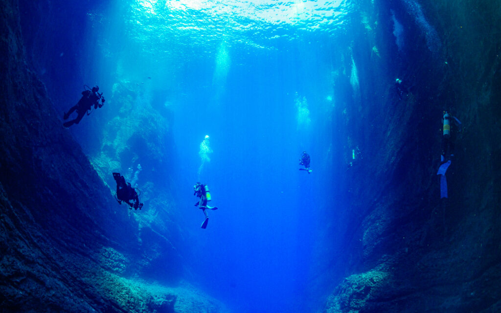Scuba divers sitting by a narrow underwater canyon, bathed in atmospheric blues