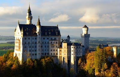 a castle with a clock on the tower of the city with Neuschwanstein Castle in the background
