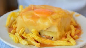 A sizzling plate with a mouthwatering Francesinha sandwich, topped with melted cheese, a fried egg, and smothered in a rich, spicy tomato sauce.