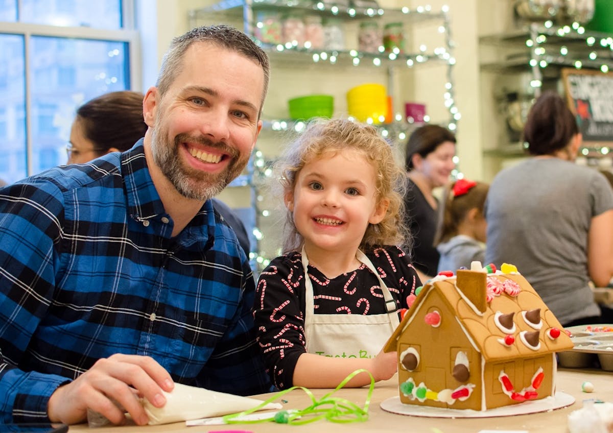 a father and daughter smiling in front of a gingerbread house
