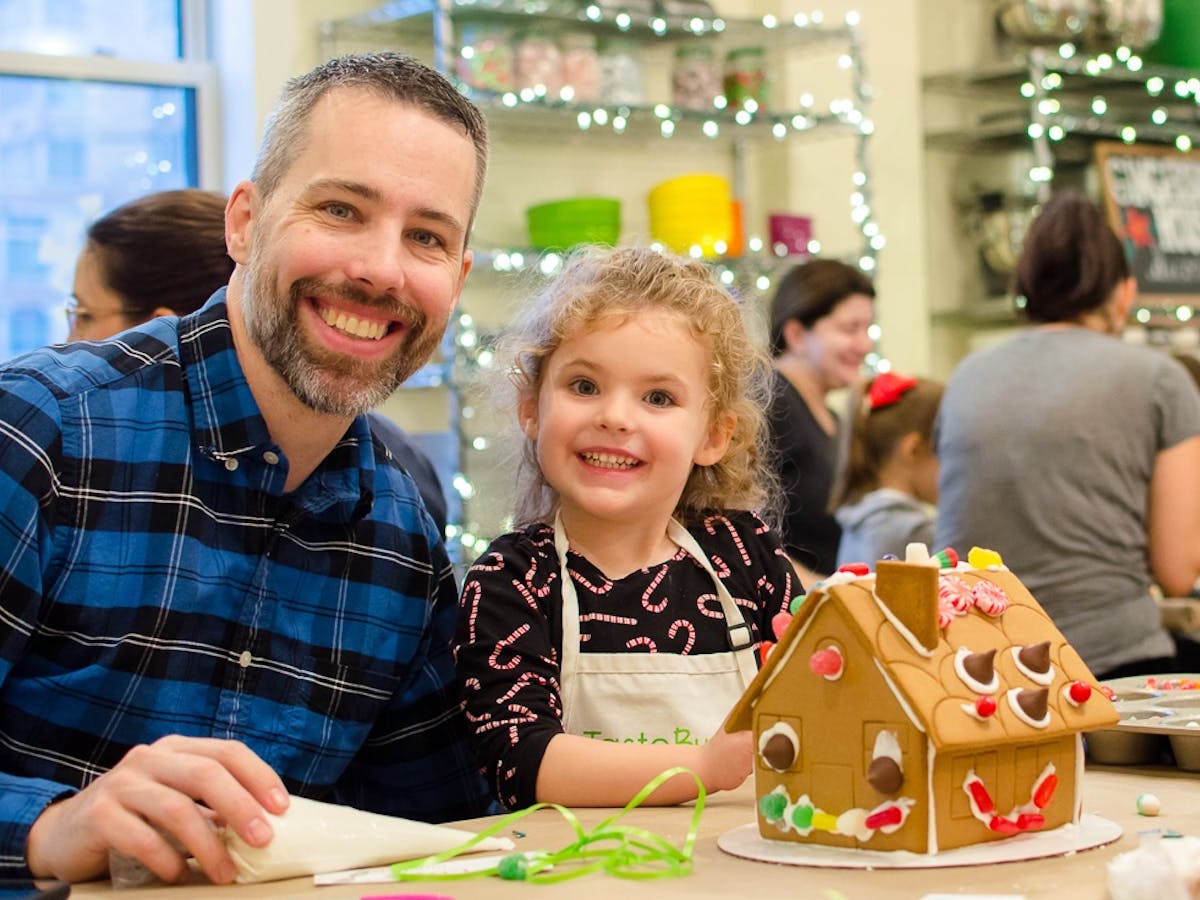 a father and daughter smiling in front of a gingerbread house