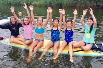 6 young girls on a paddle board with their arms raised towards the sky