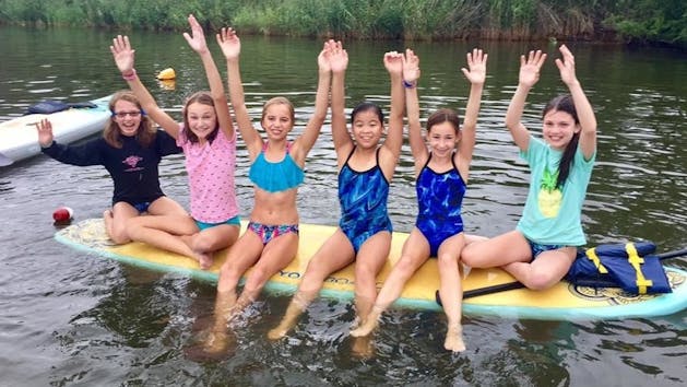6 young girls on a paddle board with their arms raised towards the sky
